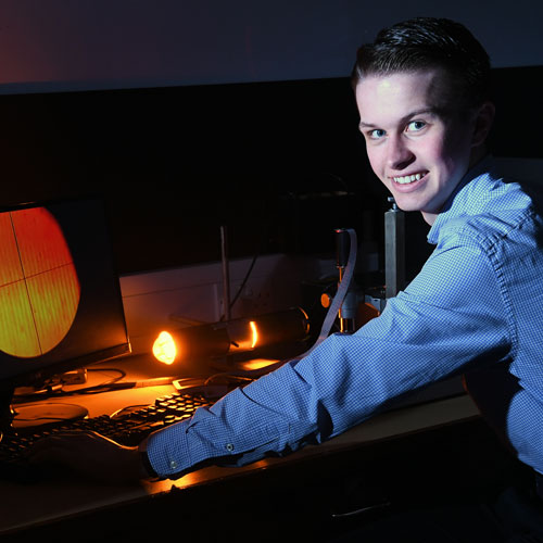 Physics with Teaching student Sam Templeton smiles at the camera while working at a computer in a physics laboratory