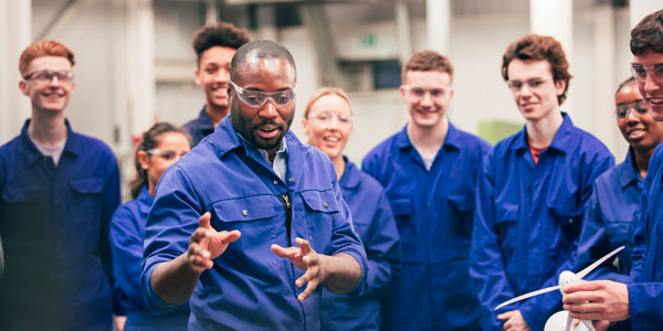 A tutor teaches his class about renewable energy in an engineering workshop. They are all wearing protective eyewear and blue coveralls.