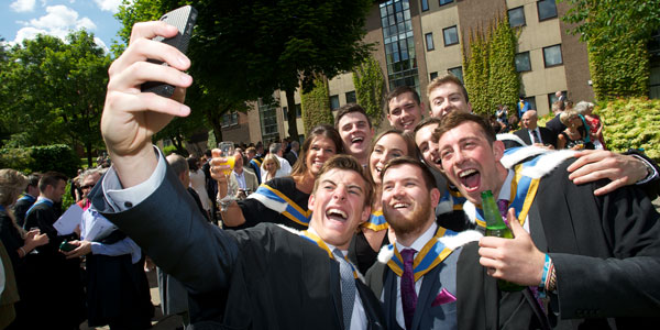 a group of students smiles together for a selfie following graduation in the Rottenrow Gardens in the sunshine