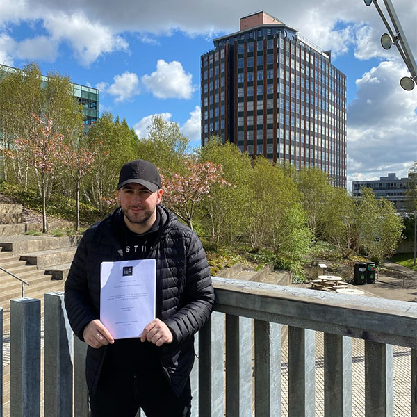 Ross McWilliam holding his dissertation with Rottenrow Gardens and Livingston Tower in the background