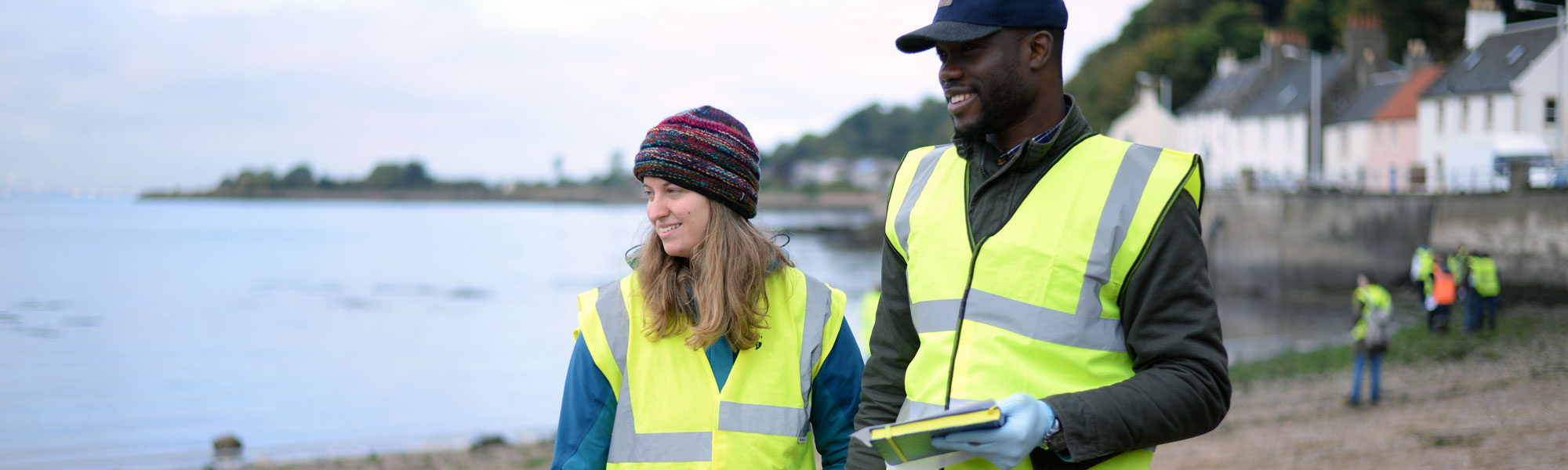 Two Civil & Environmental Engineering students wearing high vis vests on a beach in limekilns smiling and looking off camera with their classmates in the background, also wearing high vis vests, collecting samples