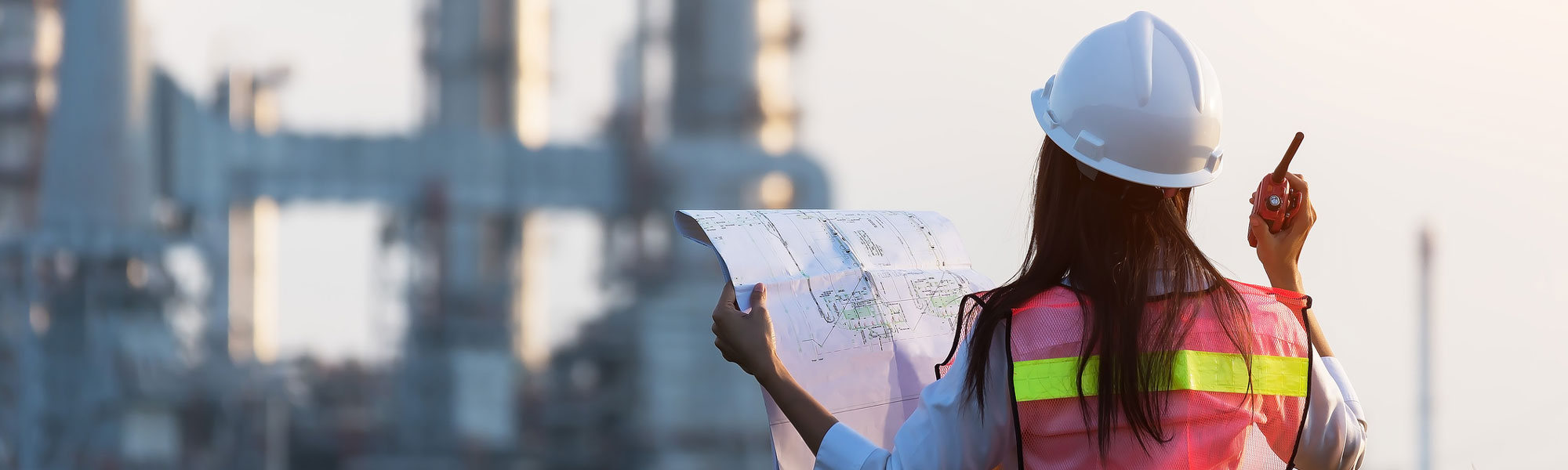 a female engineer wearing a hardhat and vest, holds a walkie talkie in one hand and a large piece of paper with plans in the other, facing towards a chemical plant