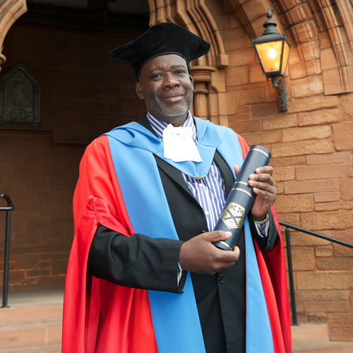 Professor John Saka smiles at the camera while holding his honorary degree parchment from the University of Strathclyde