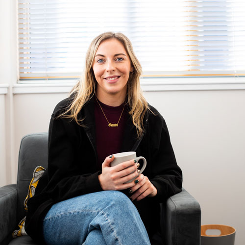 Catherine Quinn sits in an armchair with a mug in her hands, smiling at the camera