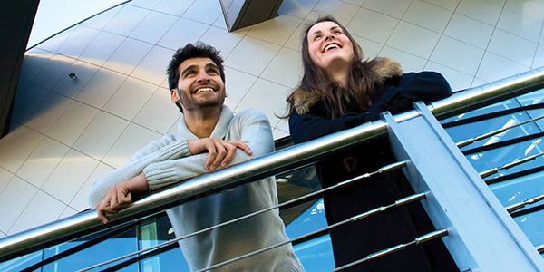 Male and female student stand on balcony on campus, 600x300