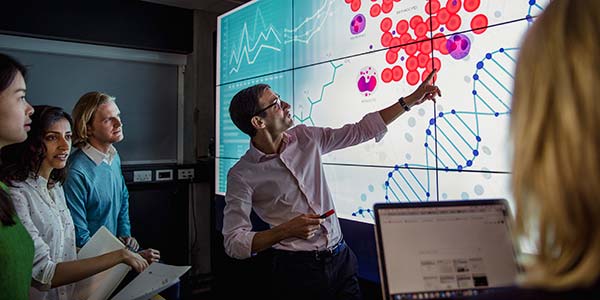 Group of business professionals room standing in front of a large data display screen with information.