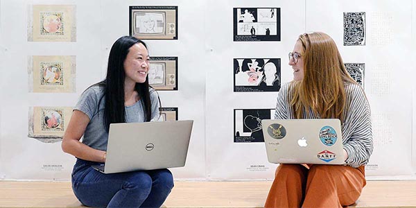 Two students sitting with their laptops. Architecture poster display on the wall behind them.