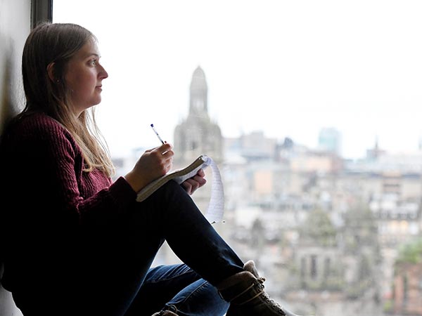 Student sitting in windowsill, writing on notepad. 