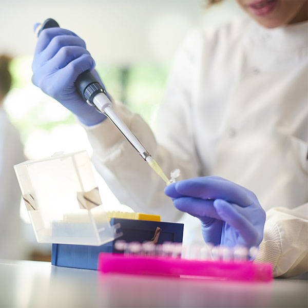 Researcher is at lab bench using a pipette to transfer samples. In the background two colleagues are placing samples in an analyser. 