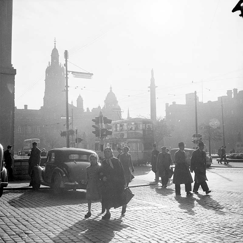 Black and white photograph of people crossing cobble streets at George Square, Glasgow, in the 1950s.