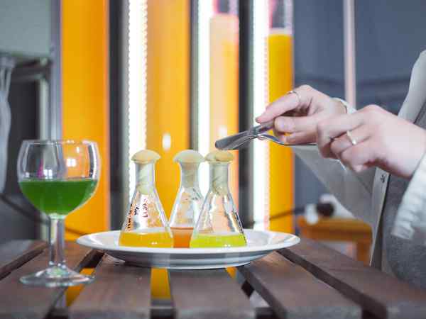 A diner uses a knife and fork to cut into a mushroom-shaped cork inside a glass beaker on a white plate with a glass of bright green liquid in a wine glass