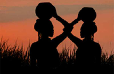 Two women carrying pots on their heads, with the sunset behind them