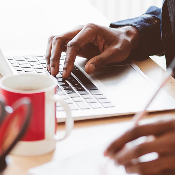 Close up of person's hands at laptop with pencil in other at a table with a mug.