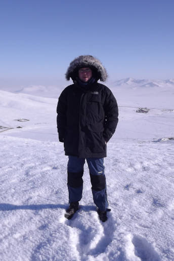 Author of photo stands in the snow with very warm jacket and some building in the far distance.