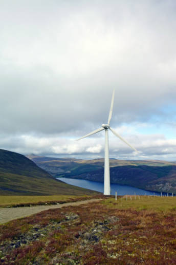 A wind turbine stands in the wild highlands, with a water body and mountains in the background.
