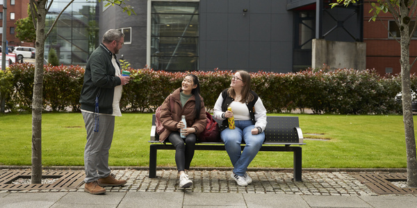 Two students on a bench, talking to another student.
