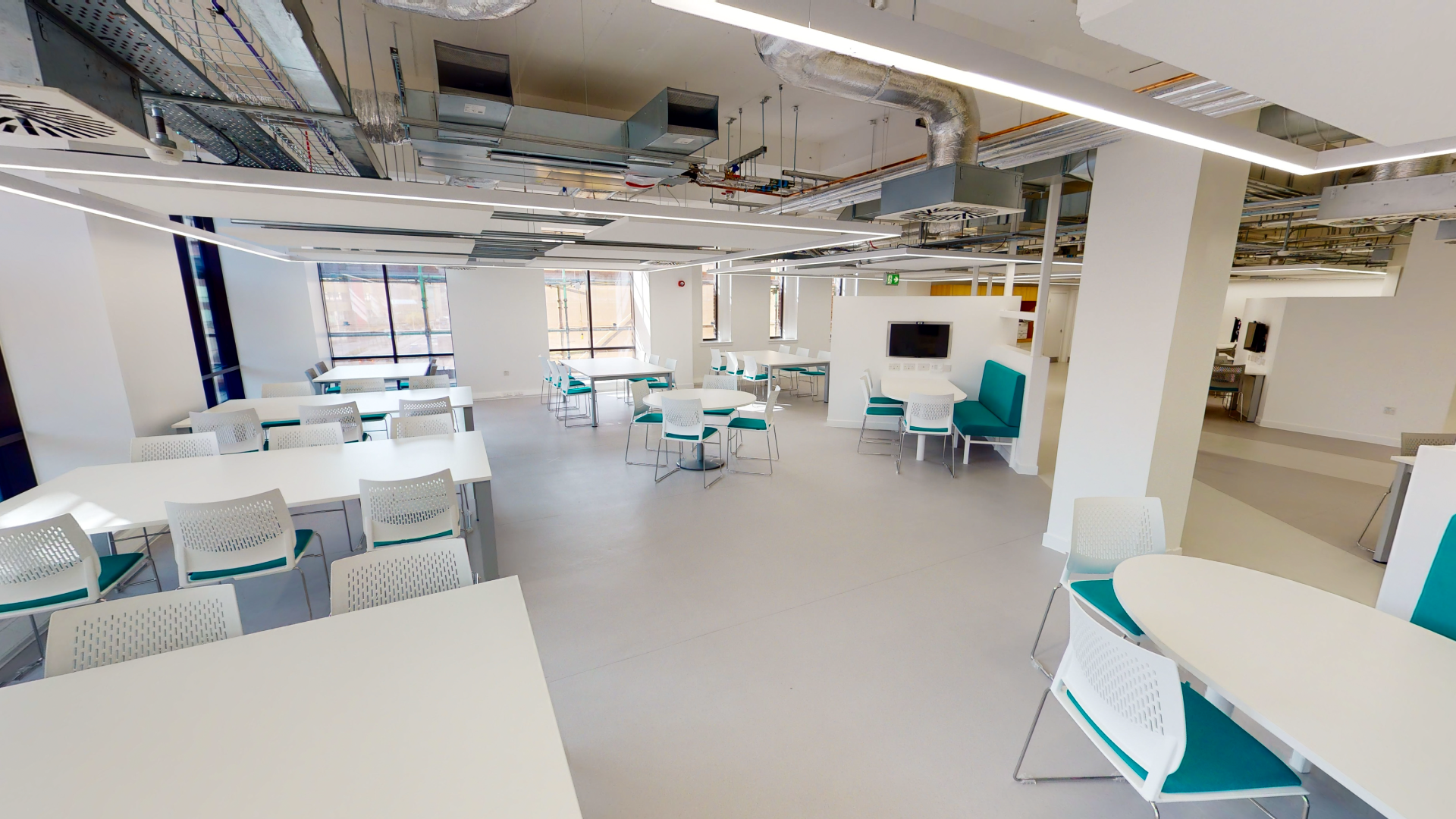 Open study area in the library, multiple big tables with chairs around them and study pods with tv screens.