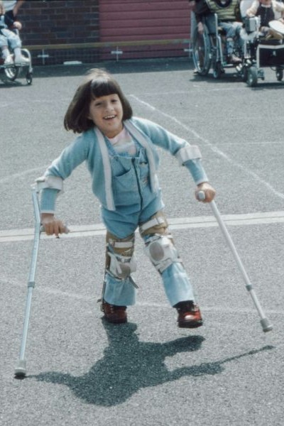 A happy disabled child uses calipers and walking sticks to walk across a playground