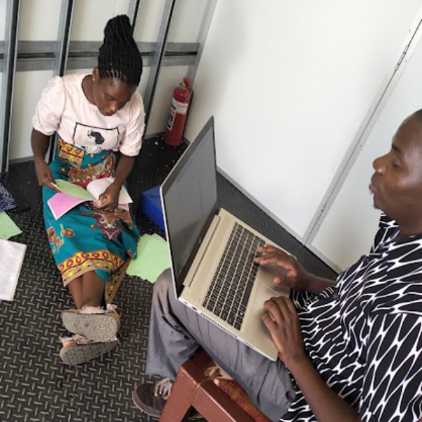 A black man sits on a chair with a laptop while a black woman sits on the floor nearby calculating receipts