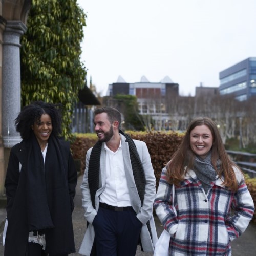 Three students smiling and walking together with university buildings in the background