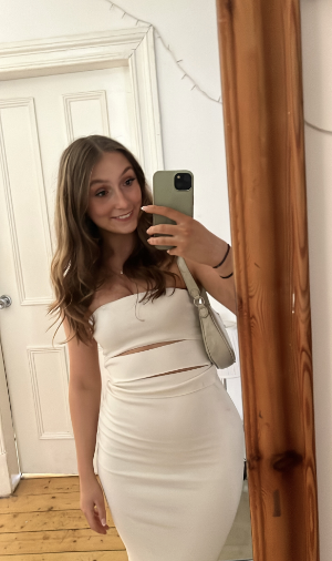 Selfie of smiling female student in front of mirror. Long brown hair and a white dress.