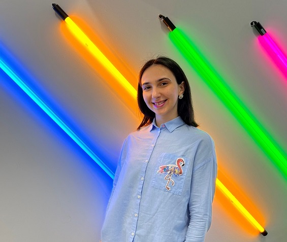Student in a blue and white pinstripe shirt with a flamingo applique on it, smiling. They're standing in front of a white wall with 4 neon tube lights attached to it. The lights are low left up to high right and are blue, yellow, green and red.
